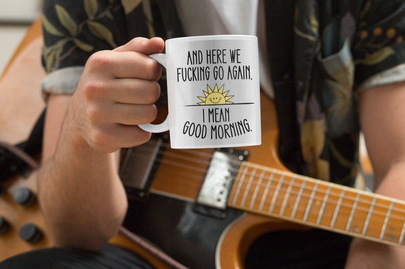 Pode incluir: Caneca de caf&eacute; branca com texto preto que diz "And here we fucking go again. I mean good morning." com um gr&aacute;fico de sol amarelo.