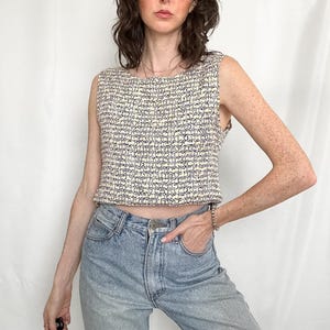 May include: A sleeveless, cropped top featuring a textured, woven knit pattern in cream and black. The garment has a round neckline and a straight hemline that sits at the waist, paired with light blue denim jeans with a classic button and zipper fly.