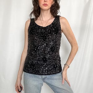 May include: A sleeveless black tank top covered in dense, shimmering black sequins arranged in a floral pattern. The garment features a rounded neckline and a straight hemline, providing a fitted silhouette that reflects light across the surface.