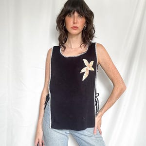 May include: A black sleeveless top featuring a white stitched trim along the edges and sides. The garment has open sides secured with black ties and a single light-colored, star-shaped appliqué on the upper left chest area of the fabric.