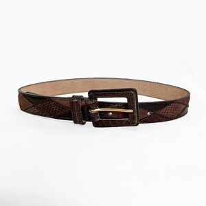 May include: A brown belt featuring a patchwork design with varying shades of dark and light brown leather. The belt has a rectangular buckle with a gold-toned metal prong and a matching brown leather-covered frame for a cohesive appearance.