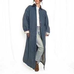 May include: A long, slate blue duster coat featuring a dark collar, a button-down front, and two large front pockets. The sleeves are rolled up to reveal a light-colored, patterned lining. The coat is worn over a white shirt and light blue jeans.