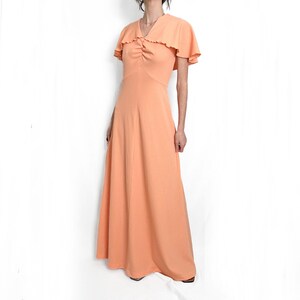 May include: A long peach-colored maxi dress with a ruffled neckline and short sleeves. The dress has a gathered bodice and a flowing skirt.