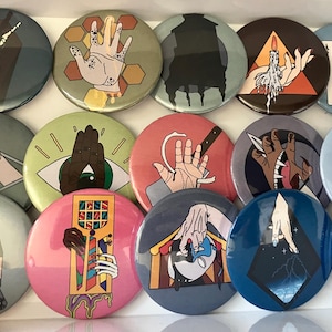 May include: A selection of round pin badges with diverse illustrations. Designs feature hands, eyes, and abstract shapes in blue, pink, green, and brown. The badges are displayed in a white box.