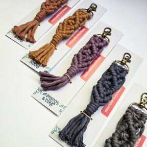 May include: Five macrame keychains with tassels in different colors, including brown, purple, and gray. Each keychain has a metal ring and is labeled "macrame keychain".