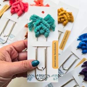 May include: A teal macrame bookmark with a gold metal paperclip. The bookmark is on a white card with the text "macrame bookmark" printed on it.