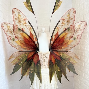 May include: A pair of fairy wings with a fall-themed color palette. The wings feature a mix of red, orange, yellow, and green hues, with a delicate, leaf-like design. The wings are attached to a white corset and a lace skirt.
