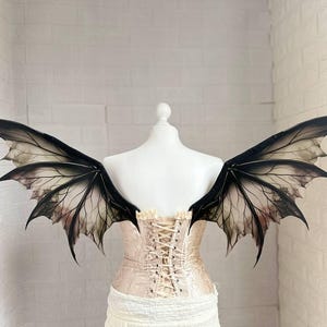 May include: Black and white fairy wings with intricate vein detailing. The wings are displayed on a mannequin wearing a cream-colored corset. The wings have a gradient effect, transitioning from black to a translucent, smoky color.