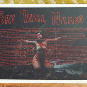 May include: A black and white print with the text "Say Their Names" in red. The print features a woman with red paint on her body and face, standing in a forest. The background is filled with handwritten names in red.