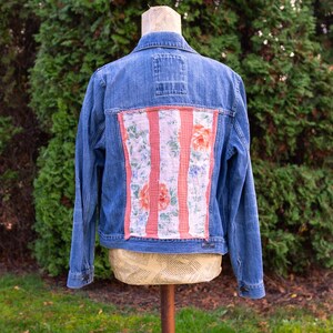 May include: A blue denim jacket with a floral design on the back. The floral design features pink and orange flowers with green leaves on a white background. The jacket is displayed on a mannequin.