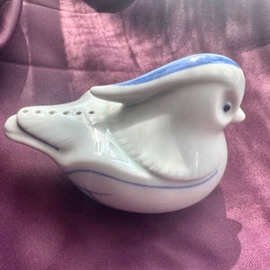 May include: A white ceramic bird-shaped salt or pepper shaker with blue accents. The bird has a small beak, a blue stripe on its head, and blue detailing on its wings and body. The shaker has several holes on its tail.