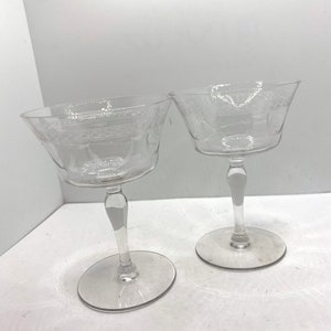 May include: Two clear glass champagne coupes with etched designs. The coupes have a delicate stem and a wide, shallow bowl.