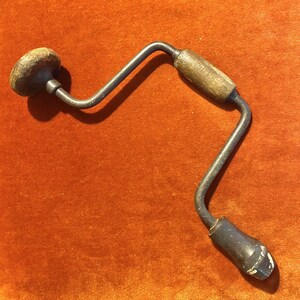 May include: A vintage hand drill with a wooden handle and a metal crank. The drill is made of metal and has a black finish. The handle is made of wood and has a brown finish.