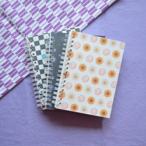 May include: A stack of three spiral bound notebooks with different patterned covers. The top notebook has a white background with orange sunflowers and yellow smiley faces. The middle notebook has a black background with white stars and a white spiral. The bottom notebook has a black and white checkered pattern.