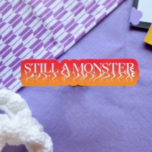 May include: A red and orange sticker with the text "STILL A MONSTER" in white letters. The text is distorted and appears to be reflected in water.
