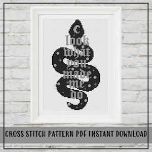 May include: A framed cross stitch pattern featuring a black snake silhouette against a white grid background. The text "look what you made me do" is written in a gothic-style font in light gray, accented with small stars and circles.