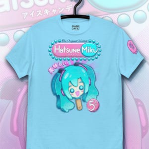 Op de afbeelding: Lichtblauw T-shirt met een afbeelding van Hatsune Miku als ijslolly. Het ontwerp bevat de tekst "Hatsune Miku" en Japanse tekst. Het shirt hangt aan een zwarte hanger.