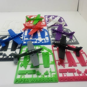 Puede incluir: Un conjunto de diez modelos de aviones Piper Warrior II impresos en 3D en varios colores, incluyendo negro, blanco, azul, rojo, verde y morado. Cada modelo está en una matriz de plástico con las palabras "Piper Warrior II" impresas en la parte inferior.