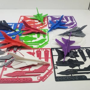 Puede incluir: Un conjunto de diez modelos de aviones impresos en 3D en varios colores, incluyendo negro, rojo, blanco, verde, azul y morado. Cada avión está ensamblado y listo para ser exhibido. Los modelos están etiquetados con los nombres "Tornado Gra", "Nakozen" y "Hakozen".