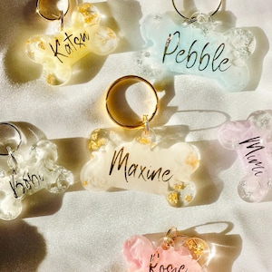 May include: Five bone-shaped dog tags with gold key rings. The tags are made of clear resin with gold flecks and have the names Katon, Pebble, Maxine, Mimi, and Rosie written in gold script.