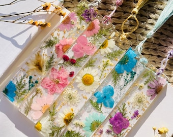 May include: Six colorful bookmarks with pressed flowers and gold leaf accents. The bookmarks are made of clear resin and have a variety of flower arrangements, including pink, blue, and white flowers.