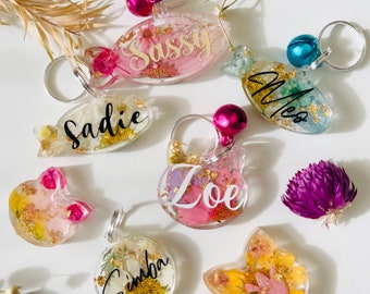 Puede incluir: Varios llaveros de resina transparente con flores secas e inclusiones de pan de oro. Cada llavero tiene un anillo plateado y una campana de color. Nombres como "Sassy", "Sadie", "Meo", "Zoe" y "Simba" están escritos.