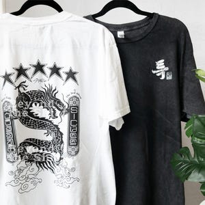 May include: Two graphic t-shirts hanging on black hangers. One is white with a black dragon design, stars, and text. The other is black with white Japanese characters. Both are short-sleeved.