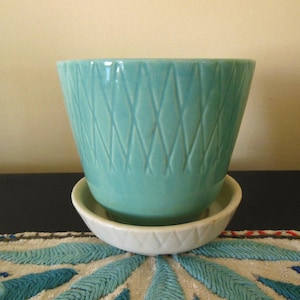 May include: A turquoise ceramic planter with a diamond pattern sits on a white saucer. The planter has a flared shape and a glossy finish. The saucer has a matching diamond pattern. The items are on a patterned surface.