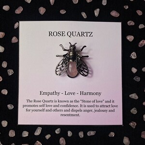 May include: A pink rose quartz bee charm on a white card with the text "ROSE QUARTZ" and "Empathy - Love - Harmony". The text below the charm reads "The Rose Quartz is known as the "Stone of love" and it promotes self love and confidence. It is used to attract love for yourself and others and dispels anger, jealousy and resentment."
