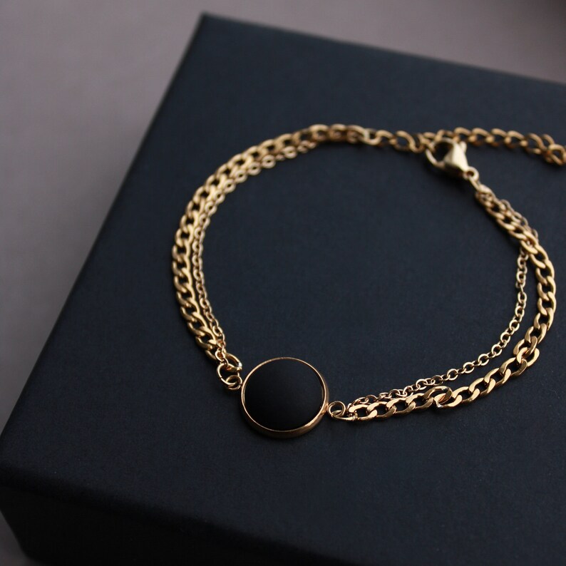 May include: A gold chain bracelet with a black circular pendant. The bracelet has two chains, one thicker than the other.