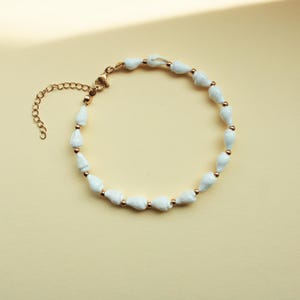 Könnte beinhalten: Ein zartes Armband mit weißen, tropfenförmigen Perlen, die mit goldfarbenen Metallakzenten aufgereiht sind. Das Armband hat einen Karabinerverschluss und eine verstellbare Kette. Der Schmuck wird auf einer hellgelben Oberfläche präsentiert.