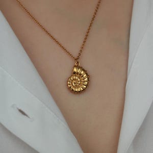 Könnte beinhalten: Eine goldfarbene Halskette mit einem Anhänger in Form eines Nautilus-Gehäuses. Der Anhänger ist aufwendig detailliert, und die Kette ist filigran. Die Halskette wird vor einem weißen Hemd präsentiert.