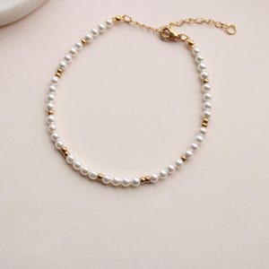 Könnte beinhalten: Ein zartes Armband mit einem sich wiederholenden Muster aus kleinen weißen, kugelförmigen Perlen und winzigen goldfarbenen Metallperlen. Das Armband verfügt über eine goldfarbene Metallkettenverlängerung mit einem Karabinerverschluss.