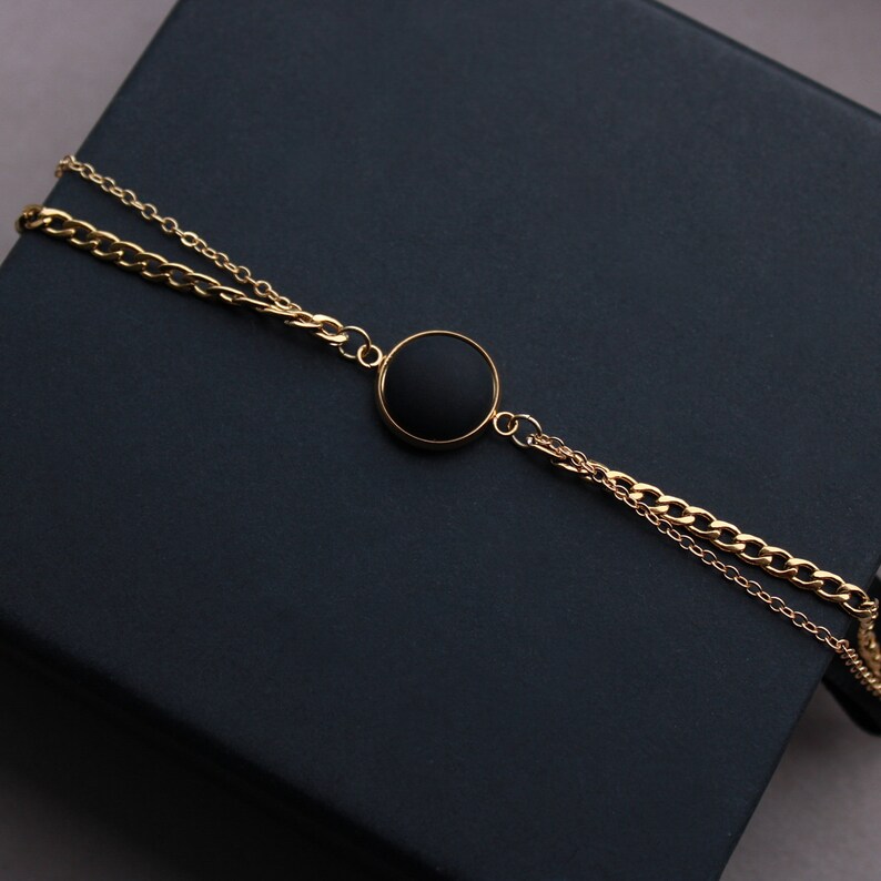May include: A gold chain bracelet with a black round pendant. The bracelet is layered with two chains.