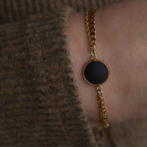 May include: A gold chain bracelet with a black circular pendant. The bracelet has two chains, one thicker than the other.
