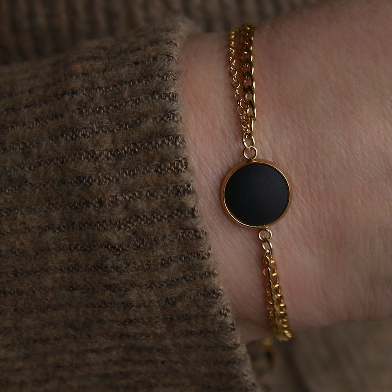 May include: A gold chain bracelet with a black circular pendant. The bracelet has two chains, one thicker than the other.