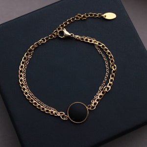 May include: A gold chain bracelet with a black circle pendant. The bracelet has two chains, one thicker than the other.