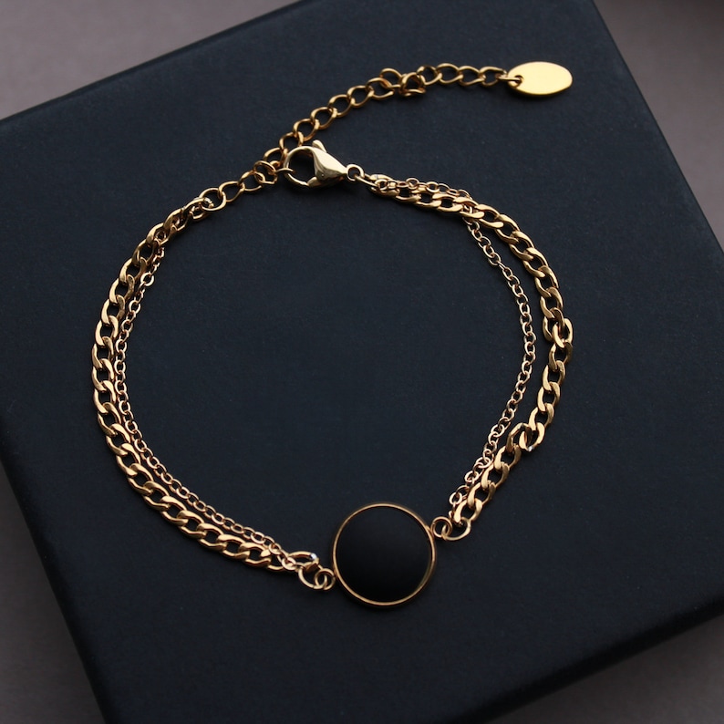 May include: A gold chain bracelet with a black circle pendant. The bracelet has two chains, one thicker than the other.