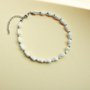 Könnte beinhalten: Weißes Muschel-Fußkettchen mit silberfarbener Kette und Akzenten. Das Fußkettchen besteht aus einer Reihe weißer Muscheln, die mit kleinen silbernen Perlen und einer Kette zur Größenanpassung aufgereiht sind. Präsentiert auf einer hellgelben Oberfläche.