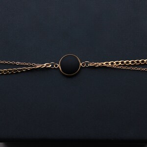 May include: A gold chain necklace with a black circular pendant. The necklace is on a black background.