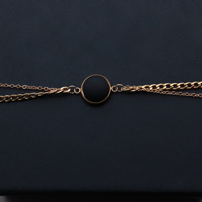 May include: A gold chain necklace with a black circular pendant. The necklace is on a black background.