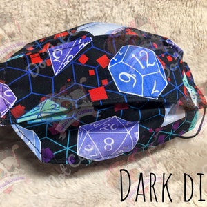 May include: A black face mask with a repeating pattern of dice in various colors, including blue, purple, and red. The dice are on a black background with a geometric pattern. The text "DARK DICE" is printed below the mask.