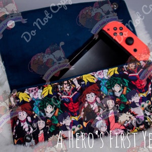 May include: A blue and black zippered pouch with a colorful print of characters from the anime "My Hero Academia". The pouch is open and a Nintendo Switch console is inside. The text "A HERO'S FIRST YEAR" is printed on the front of the pouch.