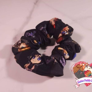 May include: Black scrunchie with a colorful anime character print. The scrunchie is made of fabric and has a gathered look. The scrunchie is on a white surface.