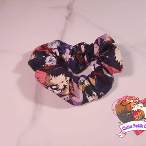 May include: A black and purple scrunchie with a print of cartoon characters from the anime My Hero Academia. The scrunchie is tied in a bow shape.