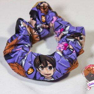 May include: Purple scrunchie with a repeating pattern of anime characters in purple, white, and brown. The scrunchie is made of fabric and has a gathered look.