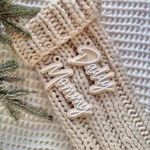 Puede incluir: Tres adornos navideños de madera con las palabras "Mommy" y "Daddy" en escritura cursiva blanca. Los adornos están colocados sobre un calcetín de punto beige. Una ramita de hoja perenne está en el fondo.