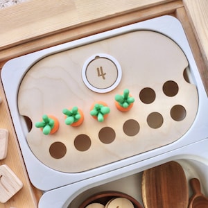 May include: Wooden educational toy set with a white tray, featuring orange carrot-shaped pieces with green tops. The tray has a wooden surface with the number 4. Additional wooden pieces and a bowl with numbered wooden discs are also visible.