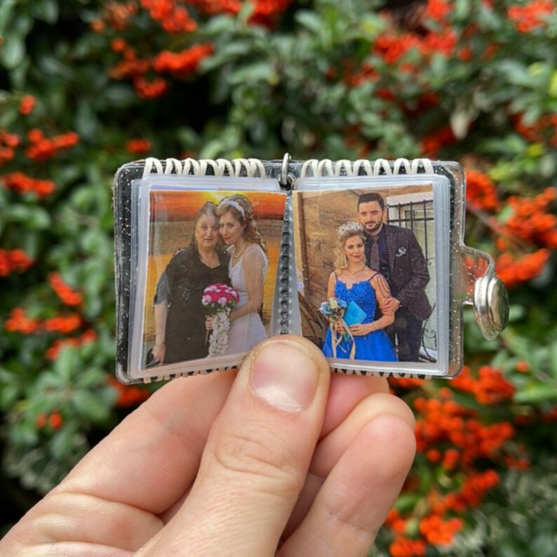 May include: A clear plastic photo album keychain with two photos. The first photo shows a woman in a white dress with a woman in a black dress. The second photo shows a man in a suit with a woman in a blue dress.