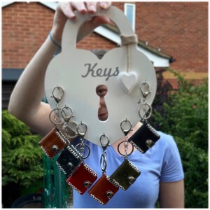 May include: A white heart-shaped key holder with the word "Keys" written on it.  The key holder has six key rings with leather key covers in various colors, including brown, black, red, and green.